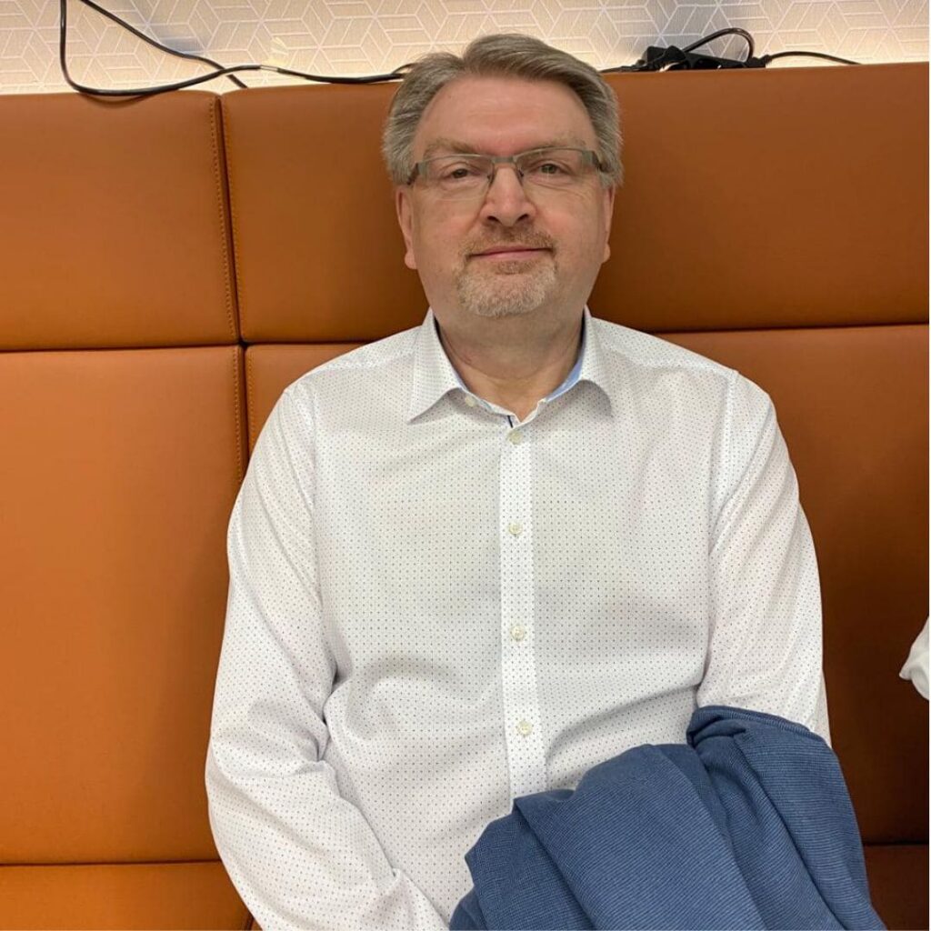Ein Mann mit Brille und weißem Hemd sitzt entspannt auf einer orangefarbenen Sitzbank und blickt freundlich in die Kamera. Das Foto vermittelt eine persönliche, offene Atmosphäre und eignet sich als Porträt für die Vorstellung einer Person oder eines Ansprechpartners.